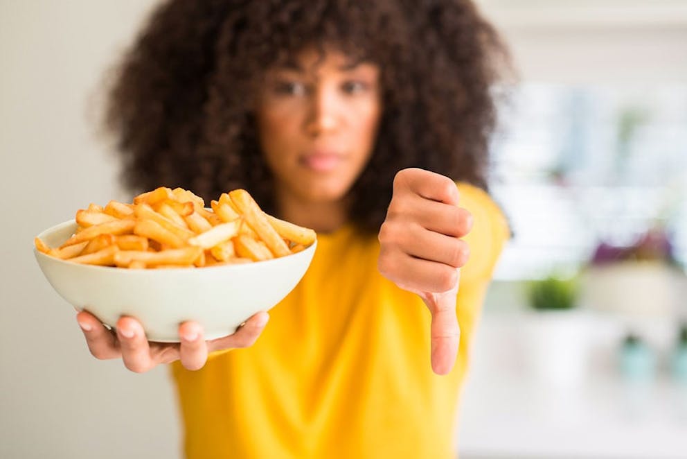 Woman holding bowl of French fries in one hand and doing thumbs down to say potatoes are bad.