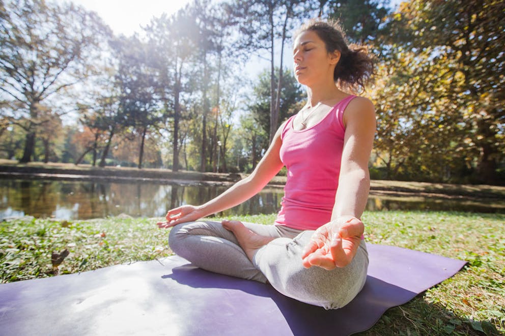 A woman practicing yoga meditation on a purple yoga mat in a park, hands on knees with eyes closed.