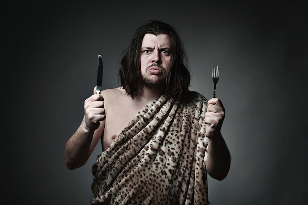 Man dressed as caveman in leopard print holds knife and fork, caveman food, what did cavemen eat.