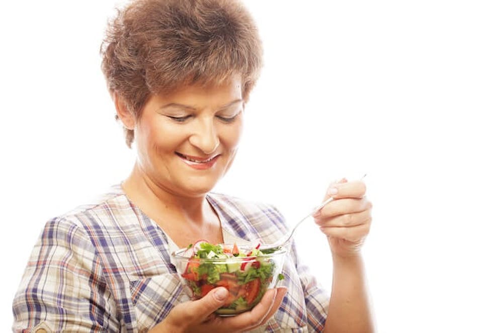A happy and smiling woman taking a bite of a healthy salad | What is Menopause…Really?