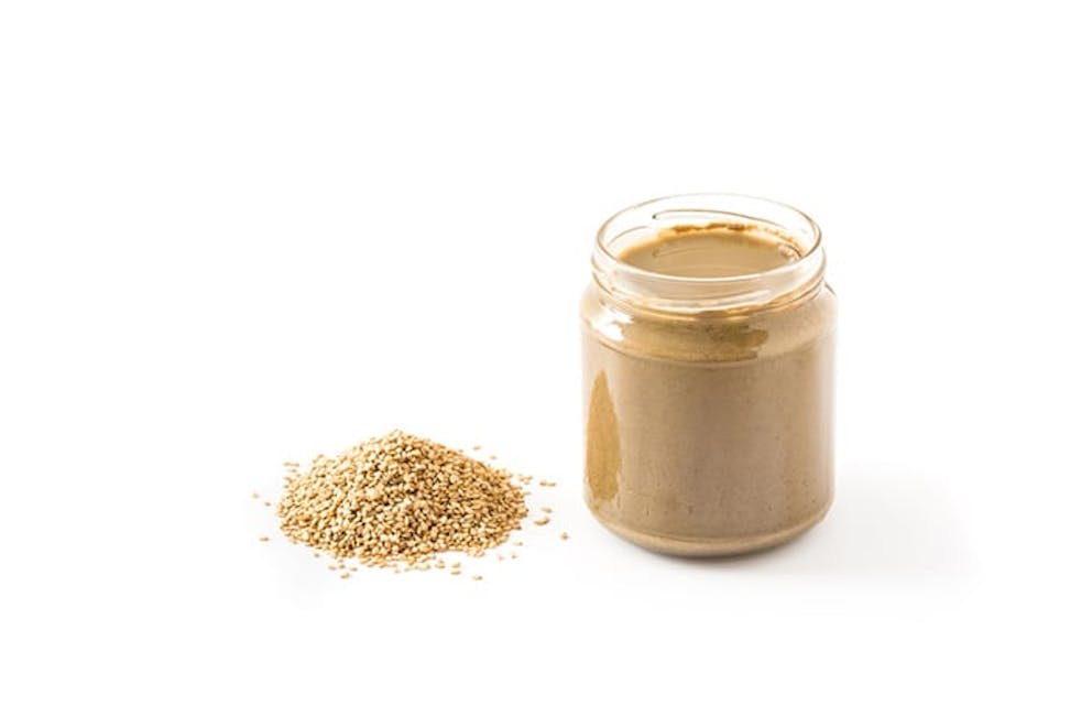 Tahini butter in glass jar next to pile of sesame seeds on white background.