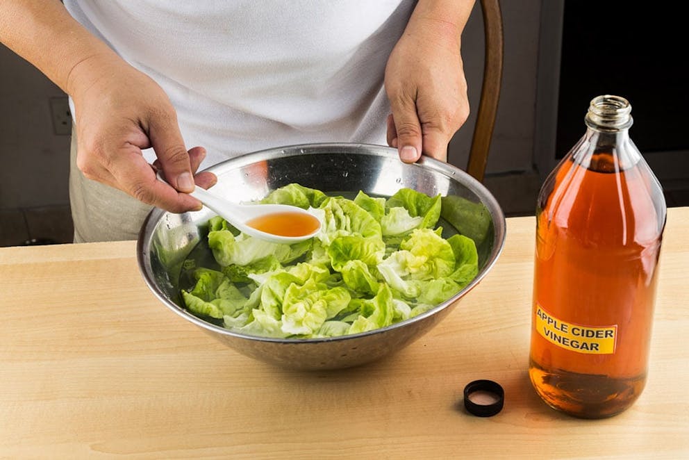 Glass bottle of apple cider vinegar with label, hands pouring it with spoon over a green salad.