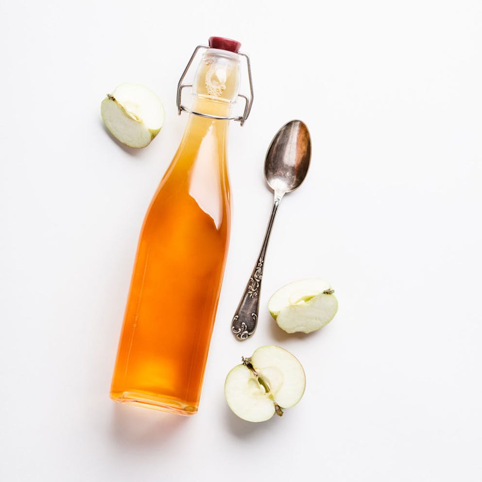 Apple cider vinegar in glass bottle with spoon and cut fresh apples nearby, ACV supplement.