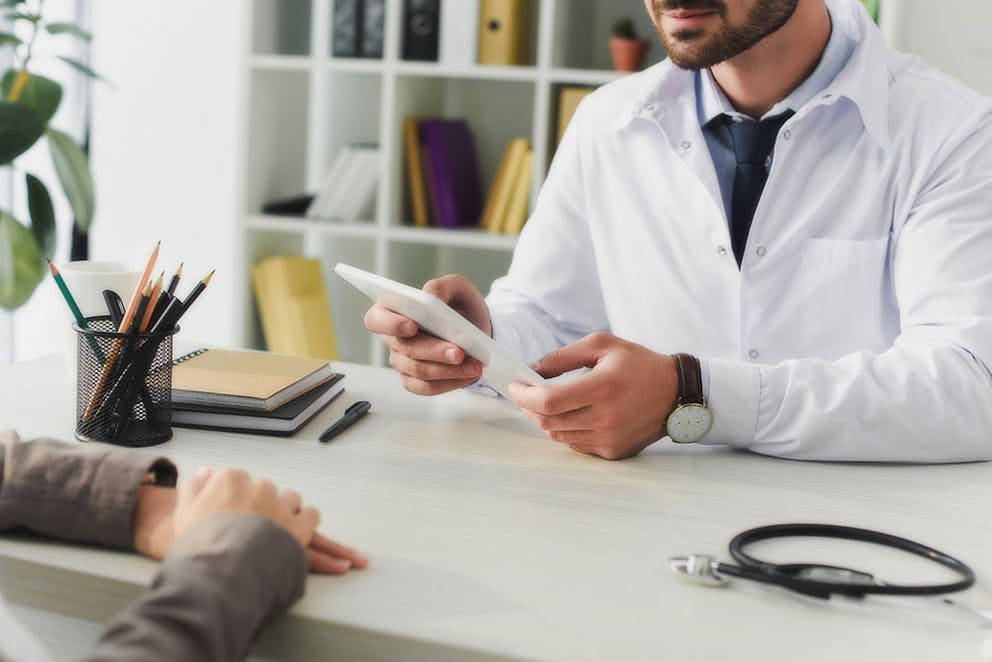 Cropped image of doctor talking with patient at a desk, using tablet to look at medical records.